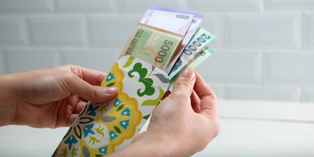 Dua tangan sedang memegang amplop kuning bermotif floral yang berisi beberapa lembar uang kertas Rupiah pecahan 5.000, 10.000, dan 20.000."