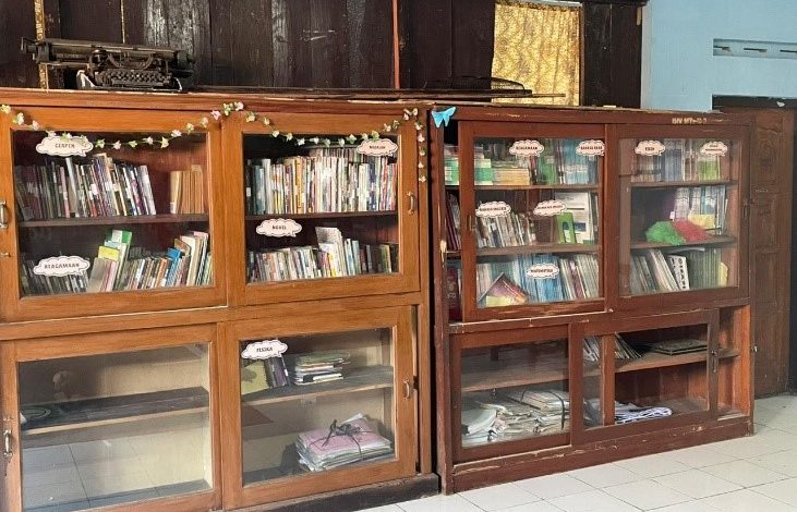 Dua lemari buku kayu besar dengan pintu kaca berisi jajaran buku sekolah. Di atas lemari terdapat mesin tik tua, dan rak-rak buku ditandai dengan label kategori seperti Fisika dan Cerpen