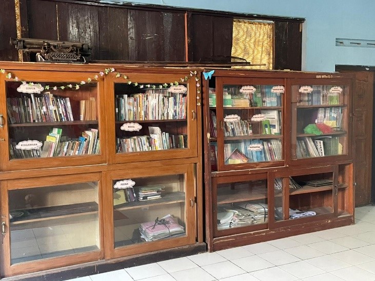 Dua lemari buku kayu besar dengan pintu kaca berisi jajaran buku sekolah. Di atas lemari terdapat mesin tik tua, dan rak-rak buku ditandai dengan label kategori seperti Fisika dan Cerpen