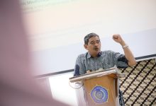 Seorang pria paruh baya mengenakan batik biru sedang berbicara di podium kayu berlogo Universitas Muhammadiyah Surakarta (UMS). Ia mengangkat tangan kirinya dengan mengepal, di latar belakang terdapat layar proyektor yang menampilkan teks presentasi