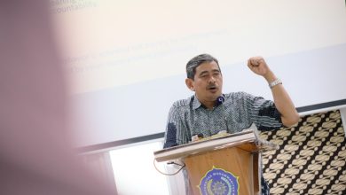 Seorang pria paruh baya mengenakan batik biru sedang berbicara di podium kayu berlogo Universitas Muhammadiyah Surakarta (UMS). Ia mengangkat tangan kirinya dengan mengepal, di latar belakang terdapat layar proyektor yang menampilkan teks presentasi