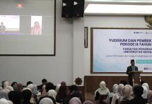 Suasana aula acara Yudisium FKIP UMS. Seorang pria berkopiah memberikan sambutan di podium, sementara layar proyektor besar di sebelah kiri menampilkan peserta melalui panggilan video daring. Puluhan mahasiswa duduk memenuhi ruangan menghadap ke depan