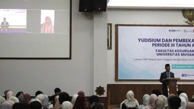 Suasana aula acara Yudisium FKIP UMS. Seorang pria berkopiah memberikan sambutan di podium, sementara layar proyektor besar di sebelah kiri menampilkan peserta melalui panggilan video daring. Puluhan mahasiswa duduk memenuhi ruangan menghadap ke depan