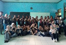Foto grup di dalam ruang kelas yang dindingnya berwarna biru muda. Sekelompok mahasiswa mengenakan almamater biru gelap berpose ceria bersama siswa sekolah yang memakai seragam pramuka. Mereka semua membentuk simbol tangan tertentu (seperti huruf T atau pose khas kelompok)