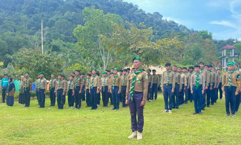 "Barisan pemuda berseragam kepanduan Hizbul Wathan (HW) sedang melaksanakan apel di lapangan rumput hijau. Mereka mengenakan kemeja cokelat, celana biru tua, serta setangan leher dan baret berwarna hijau. Latar belakang berupa perbukitan hutan yang rimbun dan beberapa tenda perkemahan