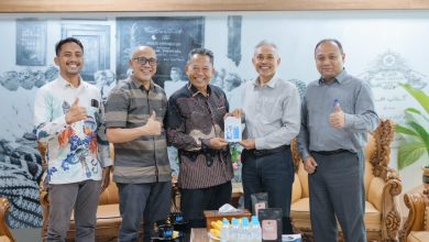 Lima pria berdiri berjejer di sebuah ruang tamu formal dengan latar belakang bertuliskan 'SURAKARTA'. Pria di tengah mengenakan batik menyerahkan souvenir kotak kecil kepada pria berbaju kemeja abu-abu terang di sebelahnya. Dua pria di sisi paling kiri dan kanan mengacungkan jempol ke arah kamera