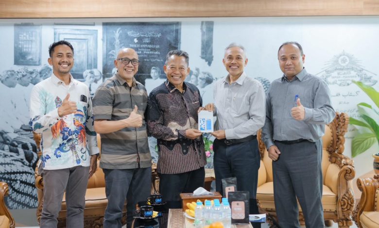 Lima pria berdiri berjejer di sebuah ruang tamu formal dengan latar belakang bertuliskan 'SURAKARTA'. Pria di tengah mengenakan batik menyerahkan souvenir kotak kecil kepada pria berbaju kemeja abu-abu terang di sebelahnya. Dua pria di sisi paling kiri dan kanan mengacungkan jempol ke arah kamera